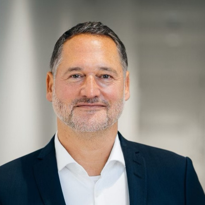 Mann mit grauem Haar und Bart in Anzug lächelt in die Kamera, unscharfer Hintergrund.