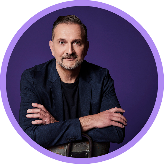 Eleganter Mann in Anzug sitzt entspannt mit verschränkten Armen vor violettem Hintergrund.