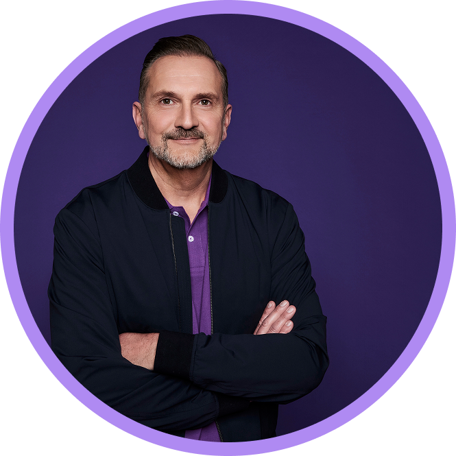 Mann mit Bart in schwarzer Jacke, lila Hemd, lächelt, verschränkte Arme vor lila Hintergrund.