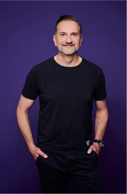 Mann in schwarzem T-Shirt vor lila Hintergrund, lächelt und hat Hände in den Hosentaschen.