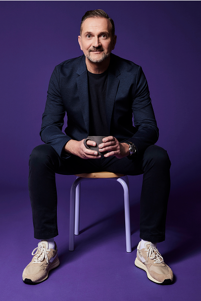 Mann in dunklem Anzug sitzt auf Stuhl, hält Tasse, lila Hintergrund.