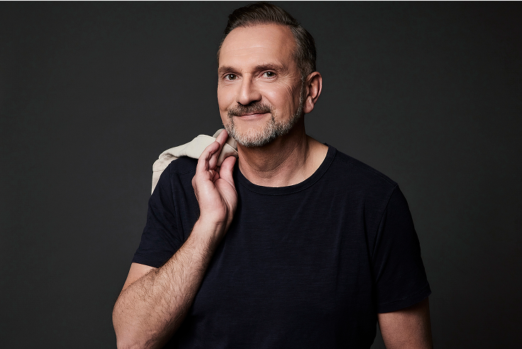 Mittelalter Mann mit Bart in dunkelblauem T-Shirt, lächelt und hält eine Jacke über der Schulter vor dunklem Hintergrund.