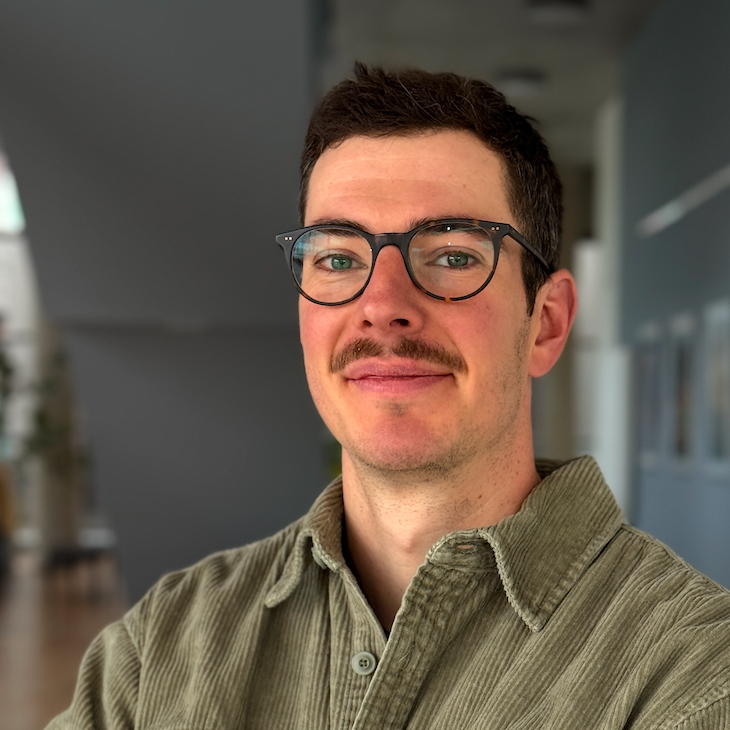 Mann mit Brille und Schnurrbart, trägt olivgrünes Hemd, lächelt leicht in moderner Innenraumumgebung.