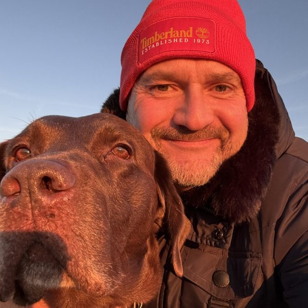 Mann in roter Mütze lächelt mit braunem Hund im Sonnenlicht, beide genießen den Moment im Freien.